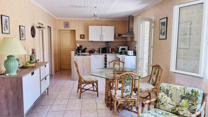 Salle à manger lumineuse avec une table, chaises, coin cuisine et décor chaleureux.