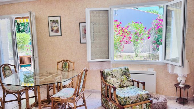 Salon lumineux avec une table en verre, des chaises et une vue sur des fleurs par la fenêtre.