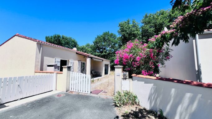 Maison avec portail, fleurs roses, et ciel bleu sur fond verdoyant.