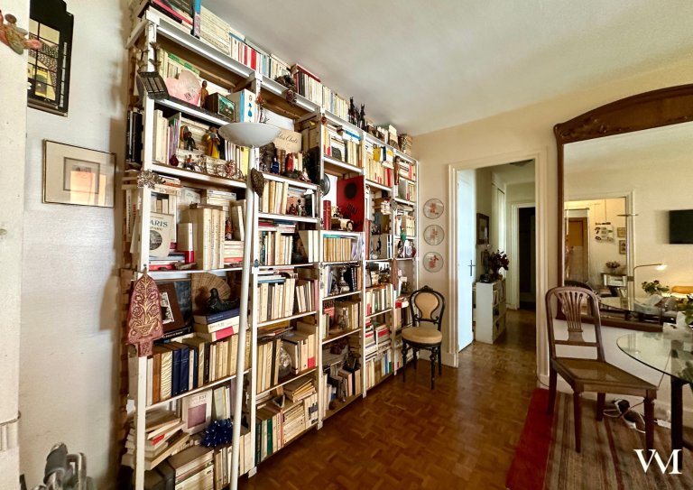 Étagère remplie de livres dans un intérieur cosy, avec des chaises et un miroir.